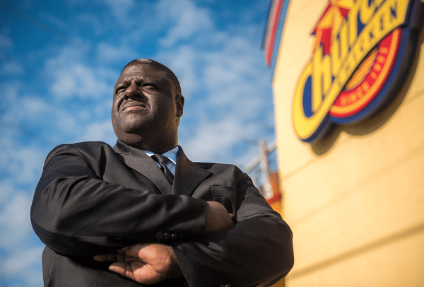 A man in a black suit and arms crossed standing in front of the Chuches Chicken building.