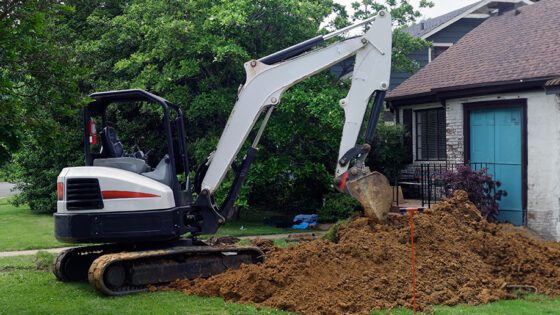 Excavator digging in a hole and creating a large dirt pile in a residential front yard.