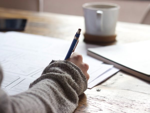 Person writing on papers with coffee and a notebook on the side.